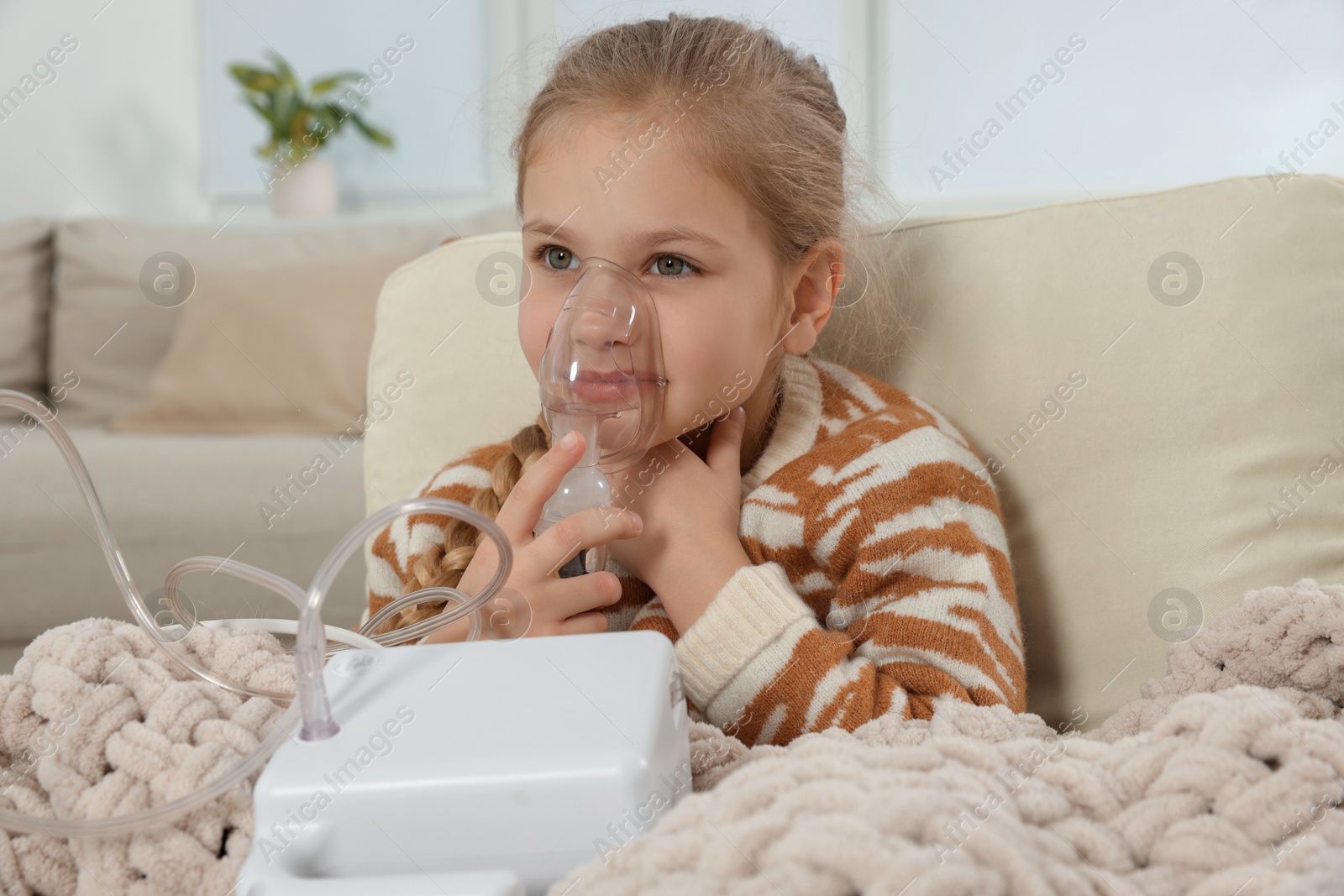 Photo of Little girl using nebulizer for inhalation in armchair at home