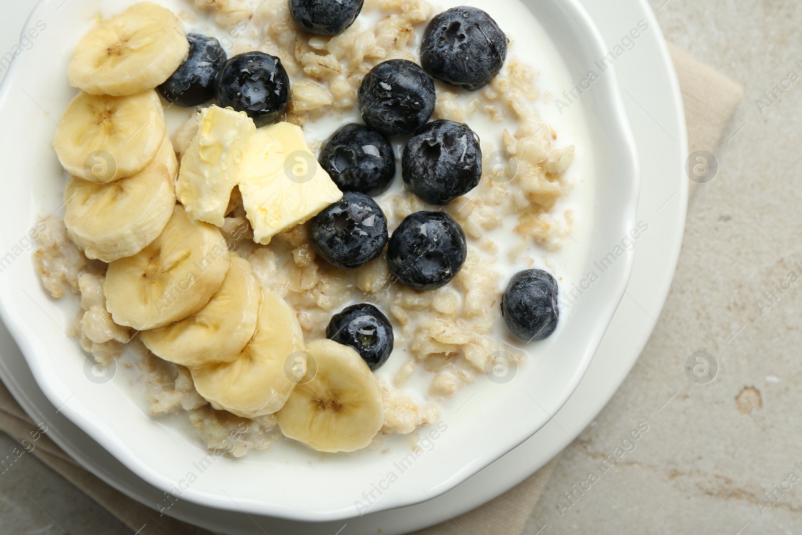 Photo of Tasty oatmeal with banana, blueberries, butter and milk served in bowl on light grey table, top view