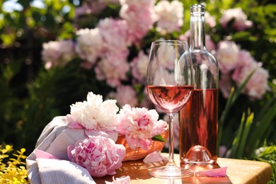 Bottle and glass of rose wine near beautiful peonies on wooden table in garden