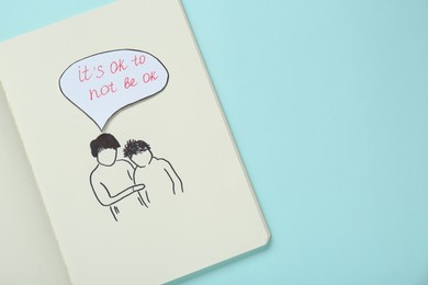Photo of Card with phrase It`s Ok to Not Be Ok, drawing of mother and her son in notebook on light turquoise background, top view. Space for text