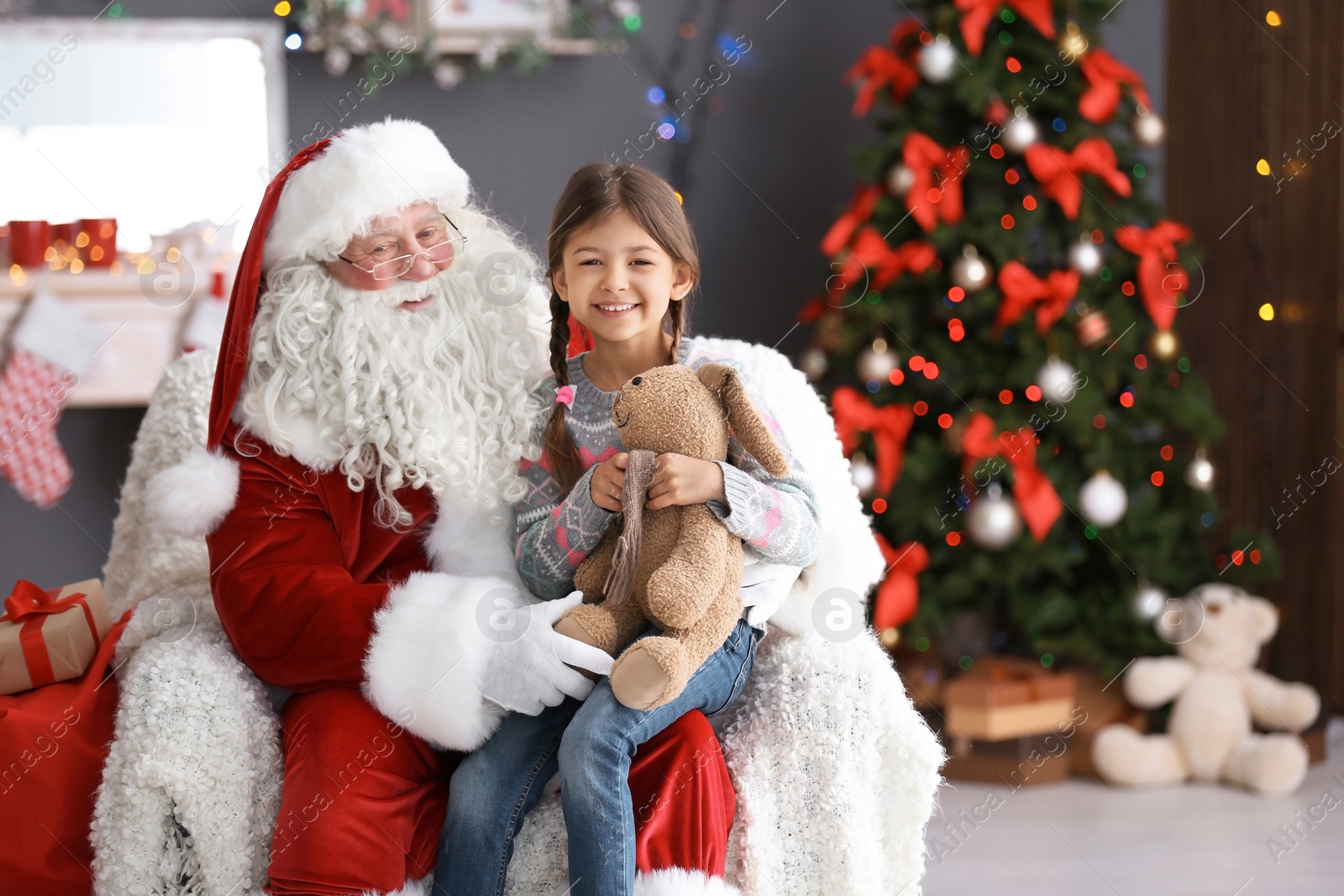 Photo of Little girl with toy bunny sitting on authentic Santa Claus' lap indoors