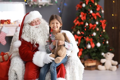 Photo of Little girl with toy bunny sitting on authentic Santa Claus' lap indoors