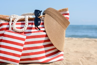 Photo of Stylish striped bag with visor cap and sunglasses on sandy beach near sea, closeup