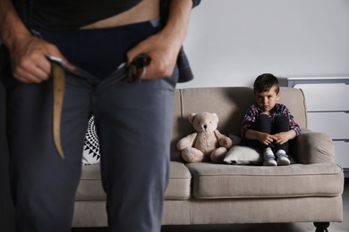 Photo of Man with unzipped pants standing near scared little boy on sofa indoors. Child in danger