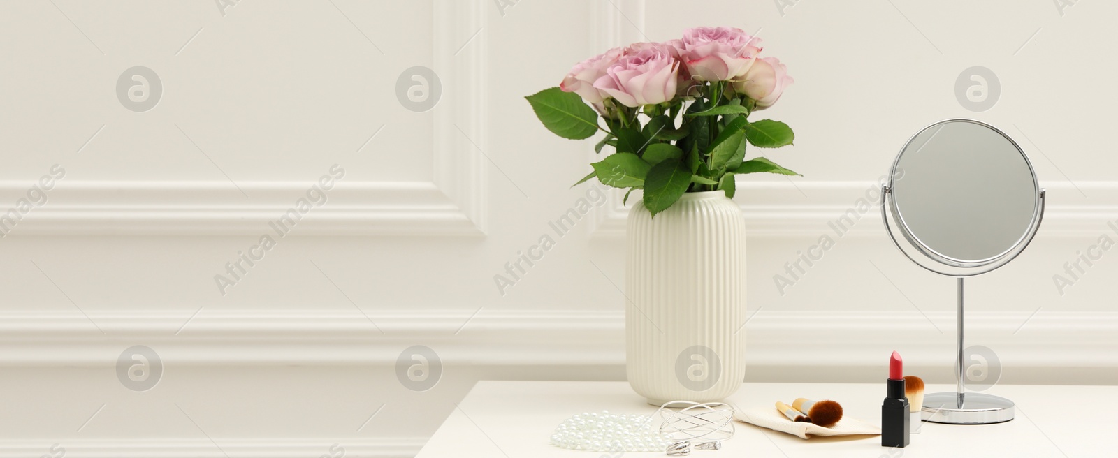 Image of Makeup room. Mirror, cosmetic products, jewelry and vase with pink roses on white dressing table indoors, space for text. Banner design