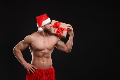 Attractive young man with muscular body holding Christmas gift box on black background, space for text