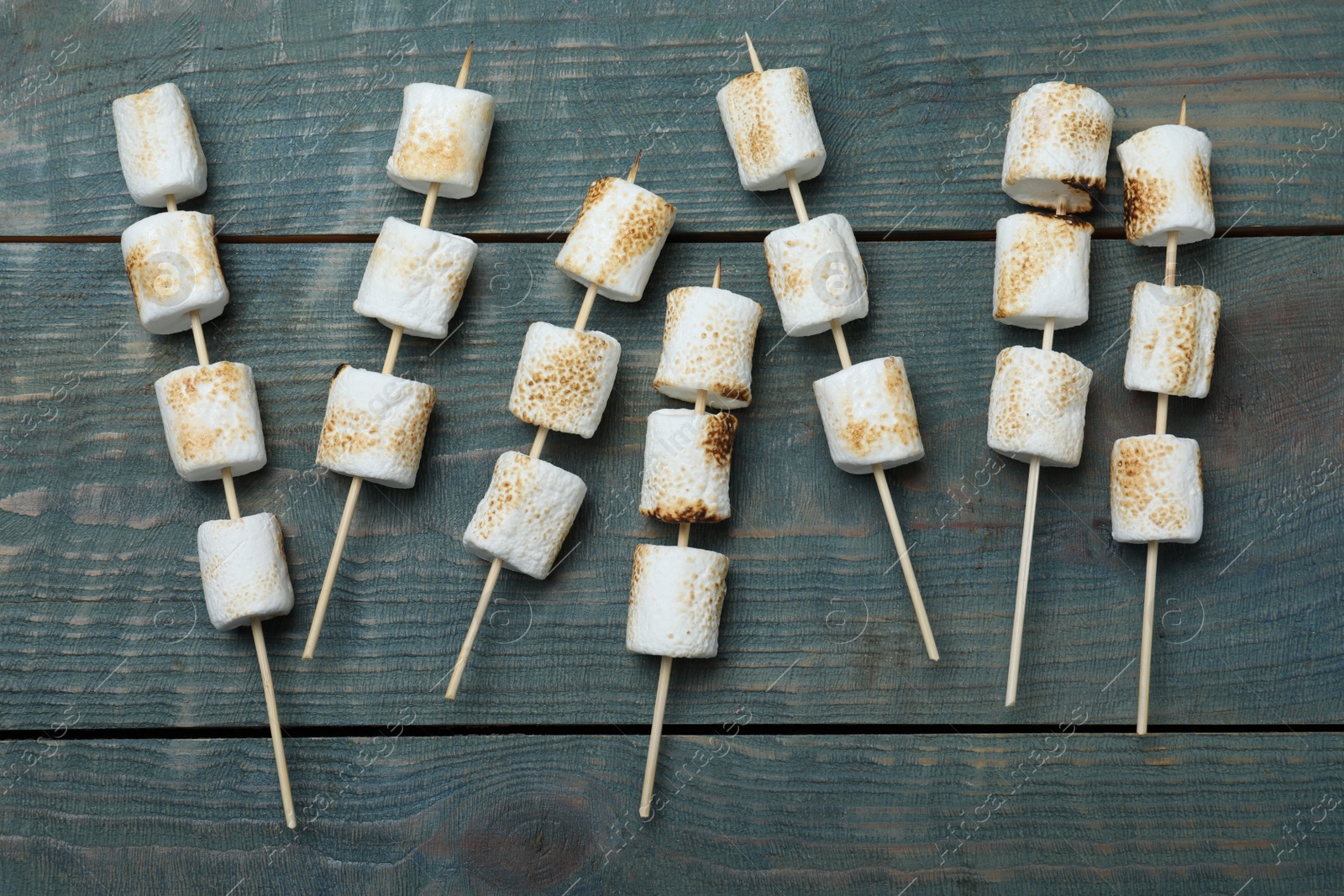 Photo of Sticks with roasted marshmallows on blue wooden table, flat lay
