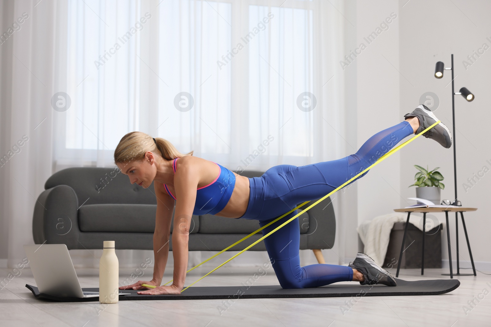 Photo of Fit woman doing exercise with fitness elastic band near laptop on mat at home