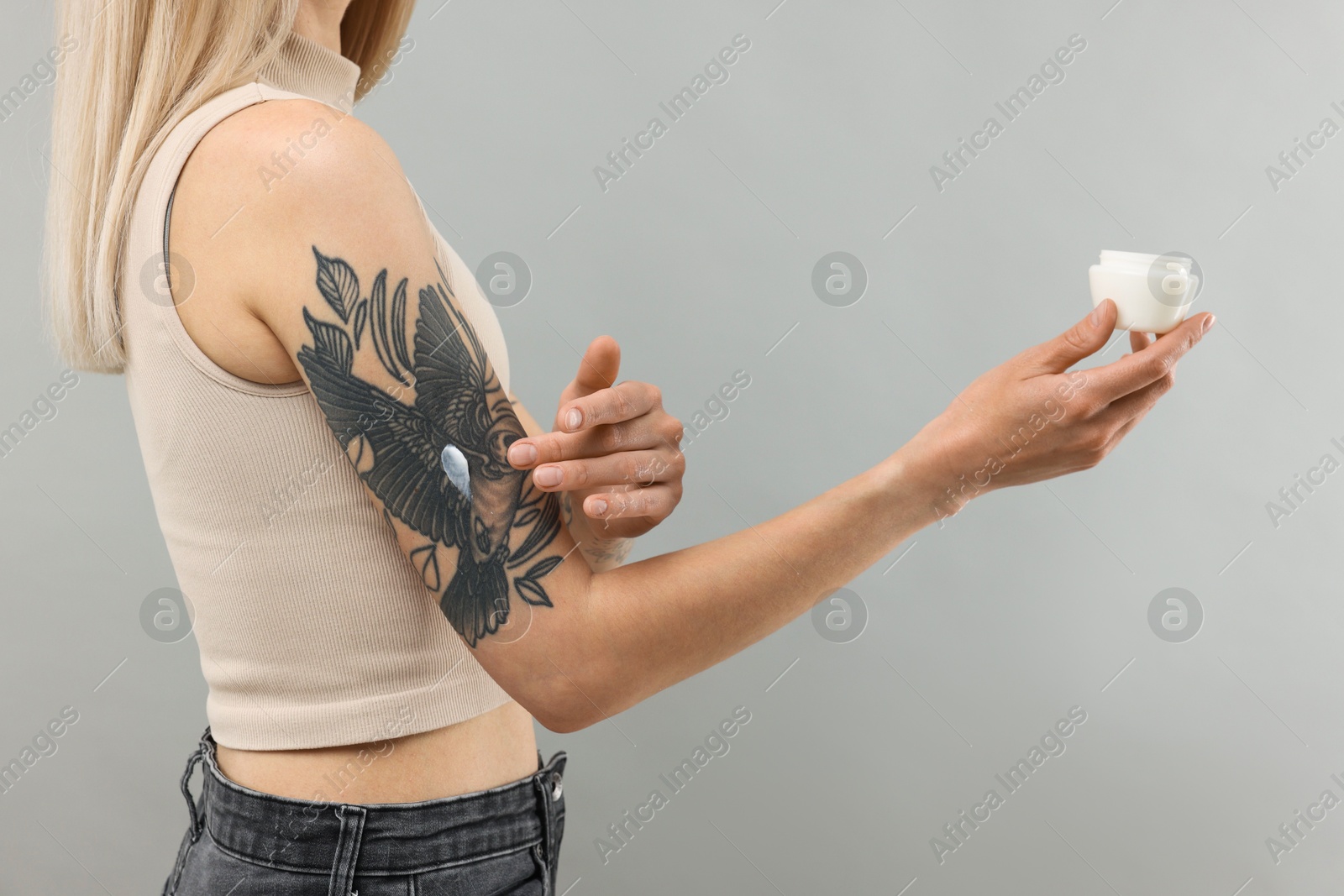 Photo of Tattooed woman applying cream onto her arm on gray background, closeup