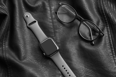 Photo of Stylish smart watch and glasses on black leather fabric, above view