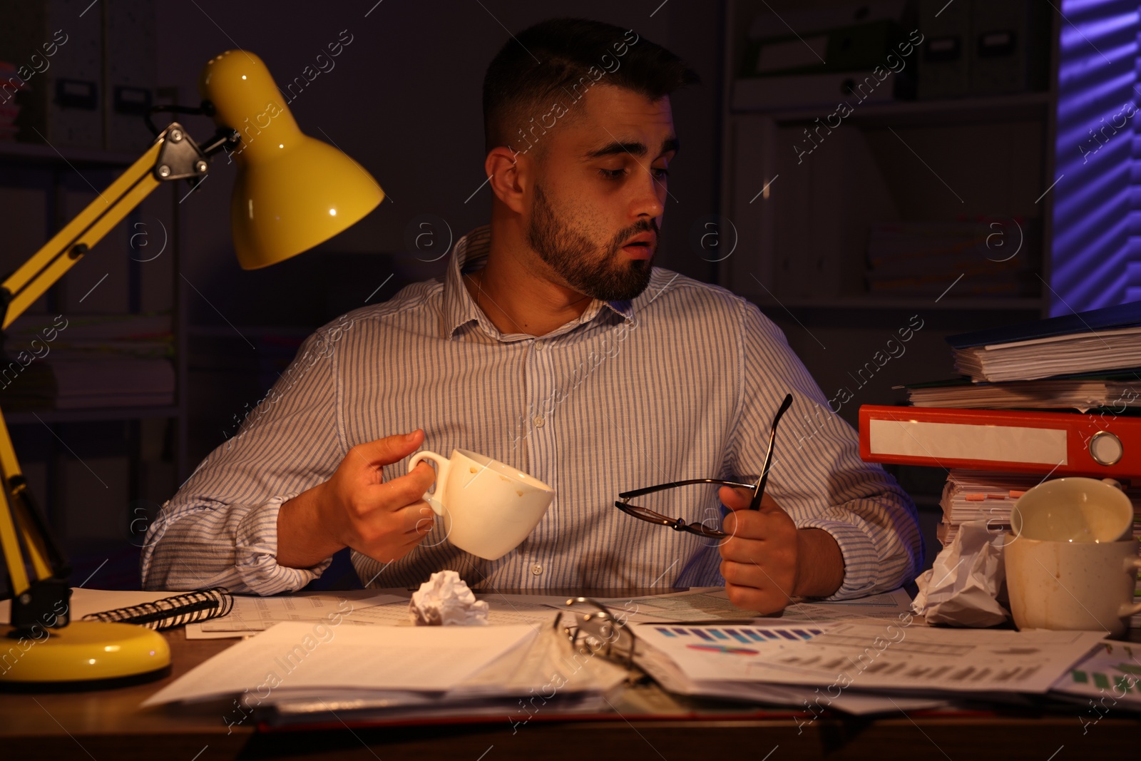 Photo of Overwhelmed man with cup at workplace in office at night