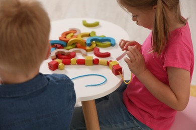 Little children playing with wooden pieces and string for threading activity at white table indoors. Developmental toys