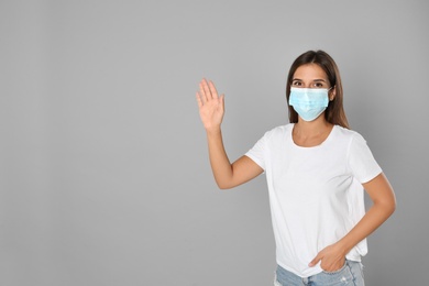 Photo of Woman in protective mask showing hello gesture on grey background, space for text. Keeping social distance during coronavirus pandemic