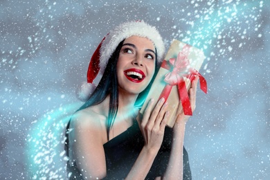 Woman in black dress and Santa hat holding Christmas gift on color background