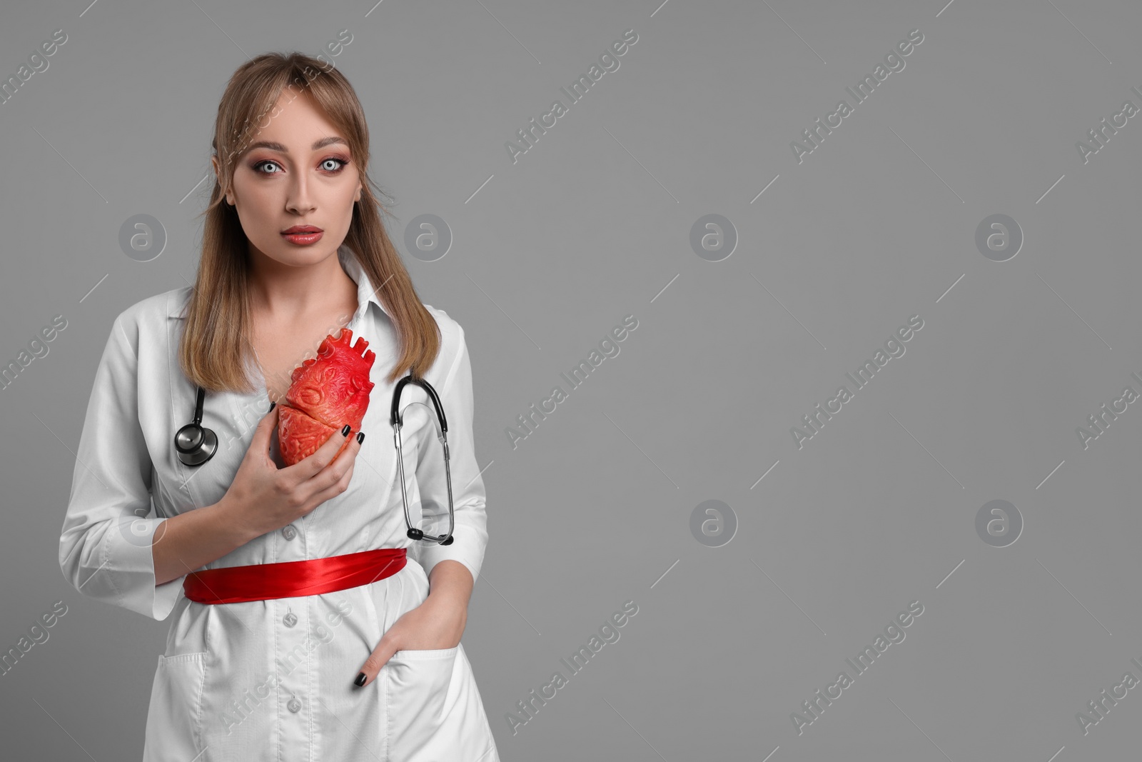 Photo of Woman in scary nurse costume with heart model on light grey background, space for text. Halloween celebration