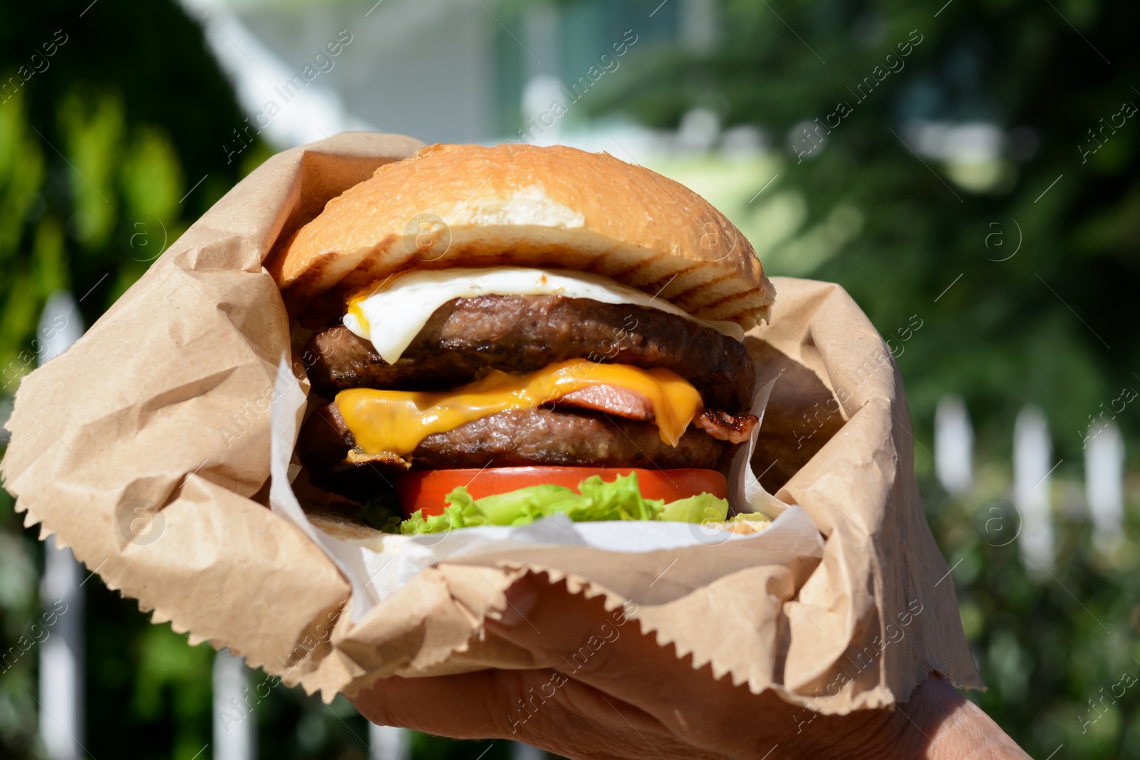 Photo of Woman holding delicious burger in paper wrap outdoors, closeup