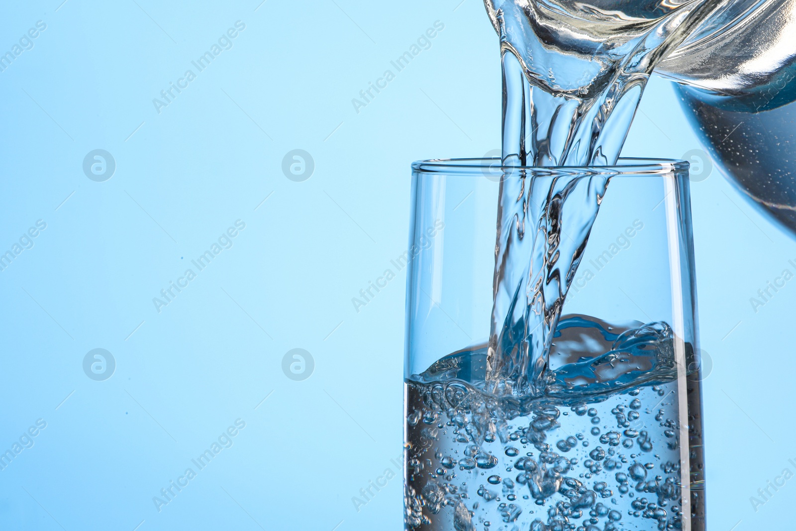 Photo of Pouring water from jug into glass on light blue background, closeup. Space for text