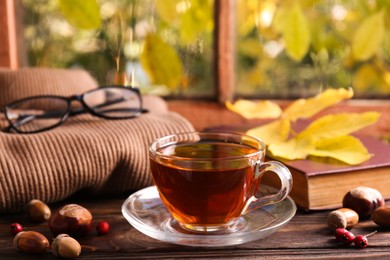 Cup of aromatic tea, acorns and soft sweater on wooden windowsill indoors. Autumn atmosphere
