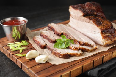 Photo of Pieces of baked pork belly served with sauce and parsley on black textured table, closeup