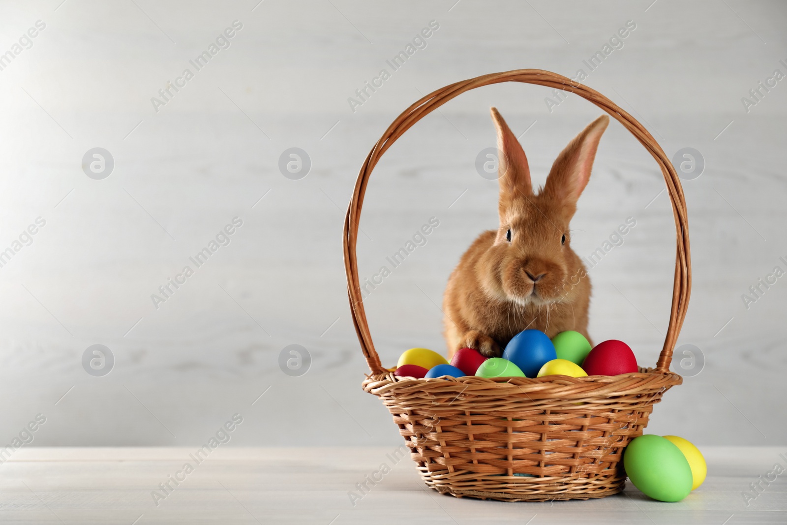 Photo of Cute bunny and basket with Easter eggs on white table. Space for text