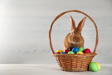 Photo of Cute bunny and basket with Easter eggs on white table. Space for text