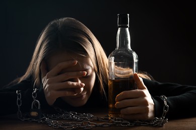 Photo of Alcohol addiction. Woman chained with bottle of whiskey at wooden table against black background