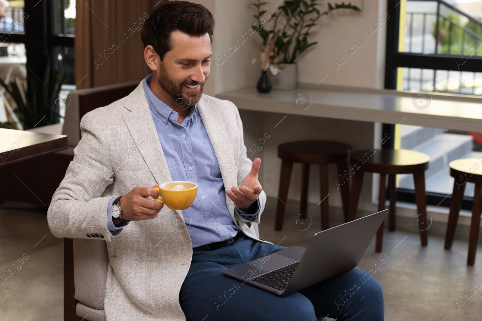 Photo of Man with cup of coffee having video chat via laptop in cafe