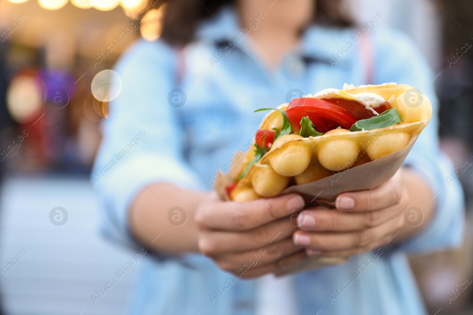 Photo of Young woman holding delicious bubble waffle with tomato and arugula outdoors, closeup