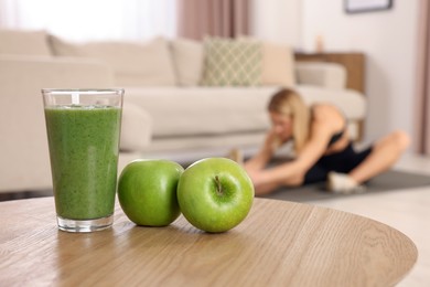 Young woman in fitness clothes doing exercise at home, focus on glass of smoothie and apples