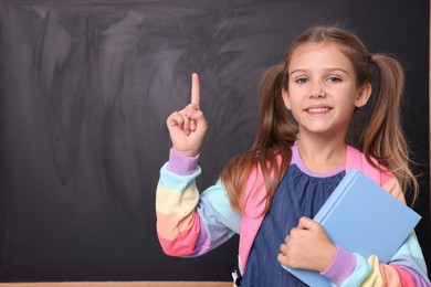Photo of Smiling schoolgirl with book pointing at something near blackboard. Space for text