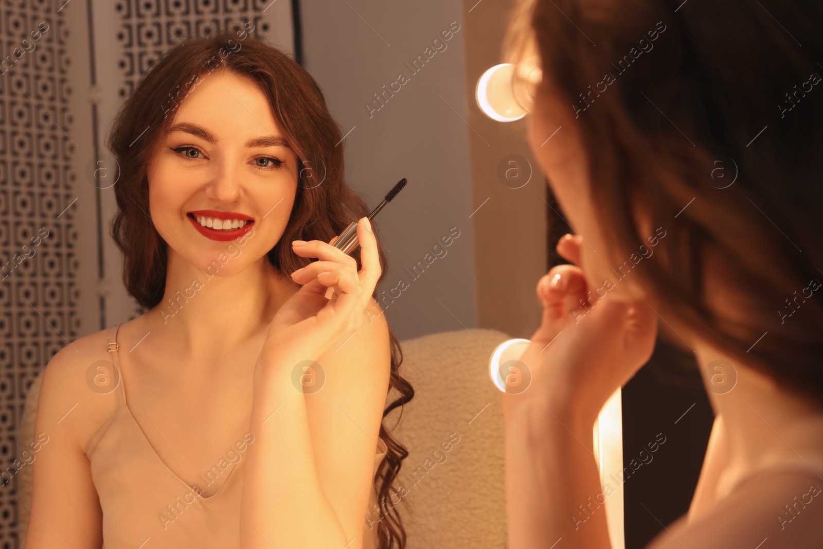 Photo of Beautiful young woman in elegant dress with mascara brush near mirror indoors