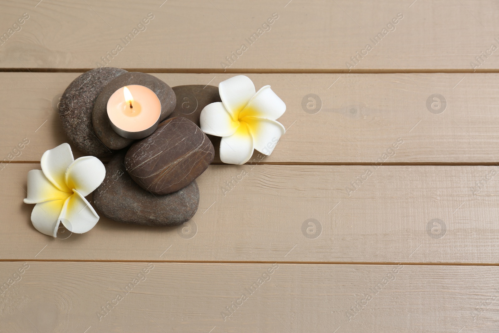 Photo of Pile of spa stones with lit candle and flowers on wooden background, space for text