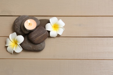 Pile of spa stones with lit candle and flowers on wooden background, space for text