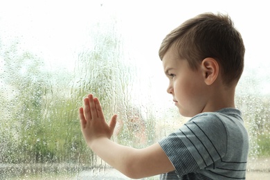 Photo of Cute little boy near window indoors, space for text. Rainy day