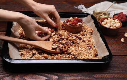 Photo of Woman putting nuts and dry fruits into tray with tasty granola at wooden table, closeup