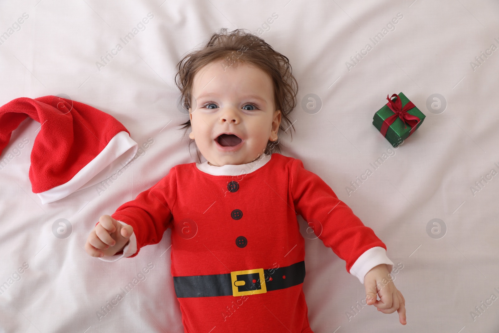 Photo of Cute baby wearing festive Christmas costume near gift box on white bedsheet, top view