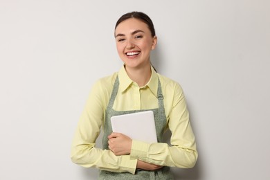 Beautiful young woman in clean apron with tablet on light grey background