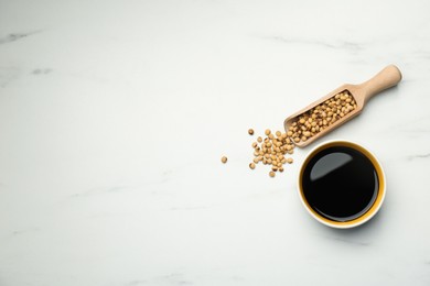 Photo of Soy sauce in bowl and scoop with soybeans on white marble table, flat lay. Space for text