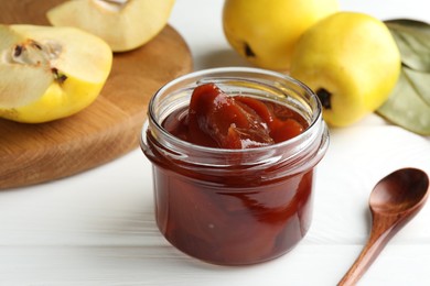 Photo of Tasty homemade quince jam in jar, spoon and fruits on white wooden table, closeup