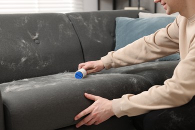 Photo of Pet shedding. Smiling man with lint roller removing dog's hair from sofa at home, closeup