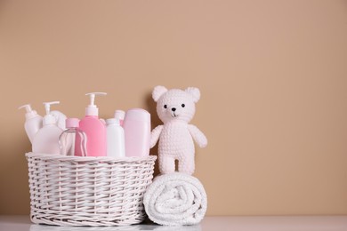 Photo of Wicker basket with baby cosmetic products, bath accessories and knitted toy bear on white table against beige background. Space for text