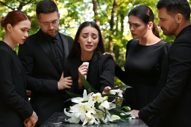 Photo of Funeral ceremony. Sad people mourning near granite tombstone with white lilies at cemetery outdoors