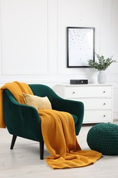 Comfortable armchair with blanket, ottoman and chest of drawers in room