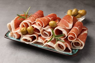Photo of Rolled slices of delicious jamon with rosemary and olives on grey table, closeup