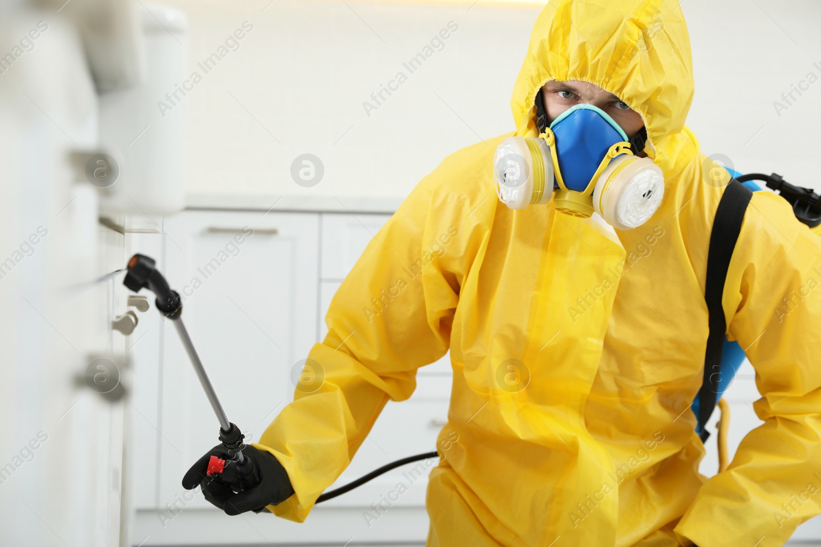 Photo of Pest control worker in protective suit spraying insecticide on furniture indoors