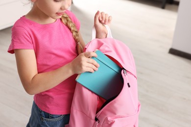 Photo of Little girl putting notebook in pink backpack at home, closeup
