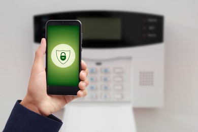 Image of Man operating home alarm system via mobile phone against white wall with security control panel, closeup. Application with illustration of padlock in shield on device screen