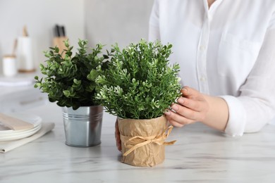 Photo of Woman near white marble table with different artificial potted herbs in kitchen, closeup