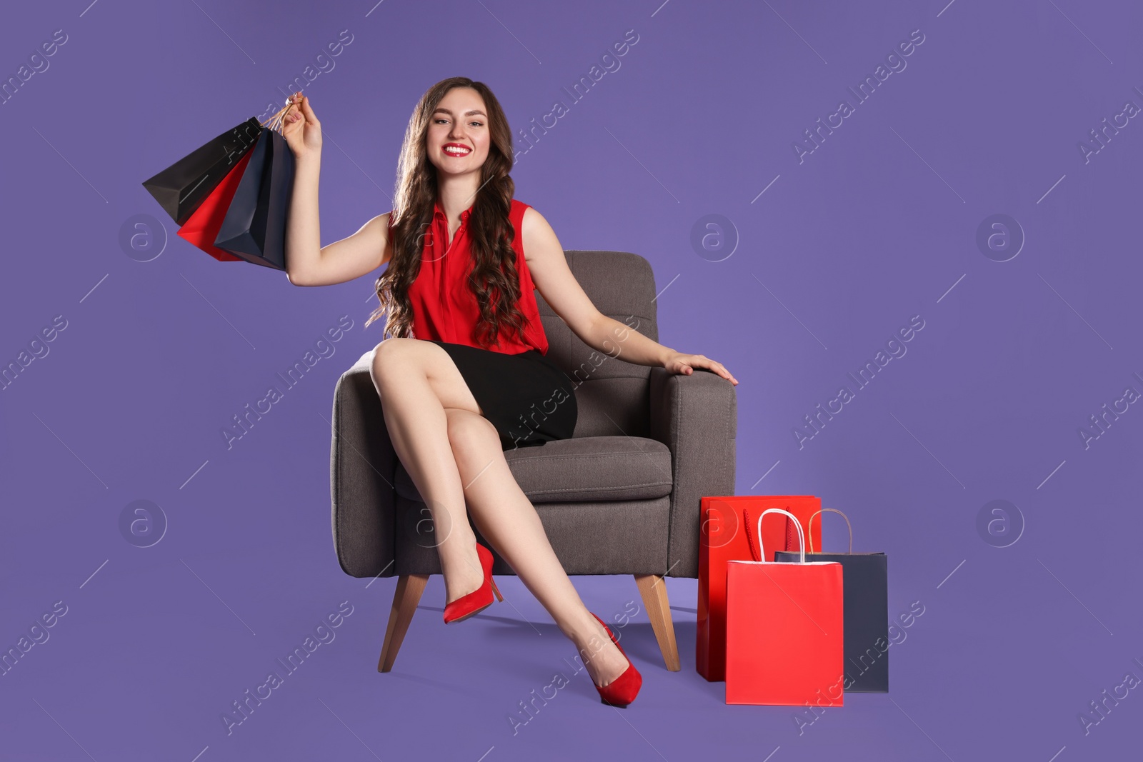 Photo of Happy woman holding paper shopping bags on armchair against purple background