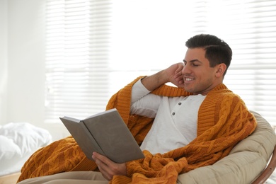 Photo of Man covered with warm orange plaid reading book at home